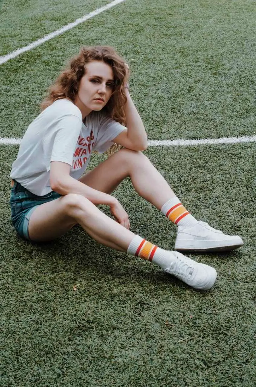 Girl sitting on football pitch in white t-shirt, touching her hair and with moody face